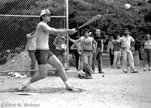 Later in the game, Jim fires a bomb to deep center field. Under the bat is John Jr., standing to his right is Danny, hands on hips is the Mailroom fellow.
